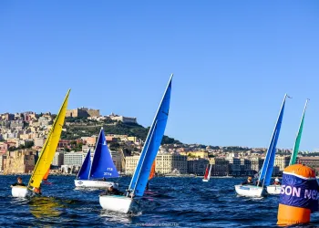 Piccole barche a vela con vele gialle, blu e verdi in regata sul mare, con la città sulla collina sullo sfondo e una boa arancione e blu