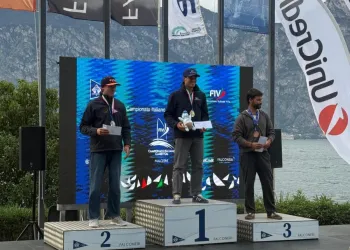 Tre uomini sul podio della premiazione davanti al lago, con bandiere e pannello blu sullo sfondo; quello al centro regge un trofeo