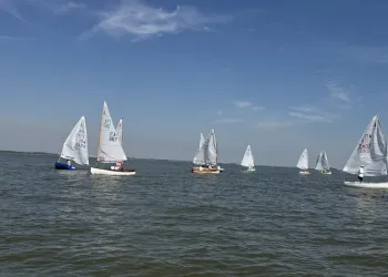 Dinghy 12' a vela sparsi su acqua calma sotto un cielo azzurro con nuvole leggere, vele bianche e linea di costa all'orizzonte