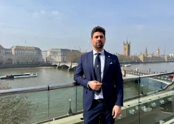Uomo in abito blu e cravatta appoggiato a una balaustra di vetro, con un fiume e un palazzo con torre sullo sfondo