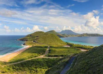 Colline verdi costiere con strada sinuosa, spiagge sabbiose e mare turchese, con un'isola montuosa sullo sfondo sotto un cielo con nuvole