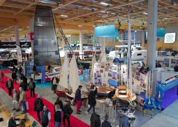 Visitatori tra gli stand di un salone nautico al coperto, con barche a vela e a motore esposte e un tappeto rosso tra i padiglioni