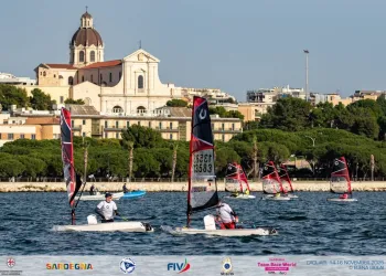 Barche OpenSkiff con vele rosse e nere in regata davanti al lungomare di Cagliari, con una chiesa a cupola sullo sfondo